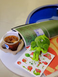 Wulibuddy Purr green Boxing Tomato Buddy plush bag  Charm & keychain accessory on a table with a cup of tea and a book