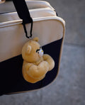 wulibuddy beige coach bear plush bag charm & keychain accessory attached to a suitcase on a gray pavement background
